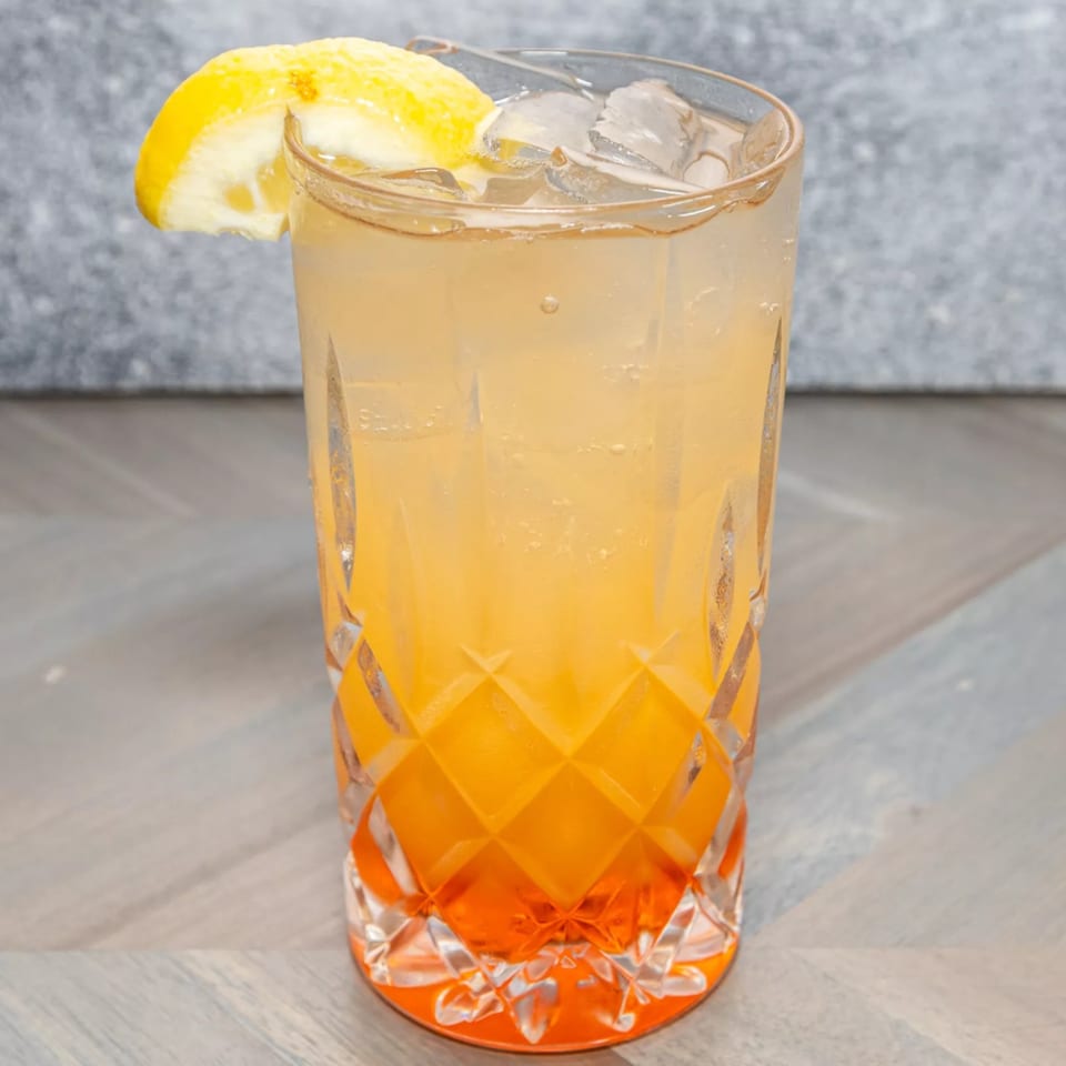 A tall, faceted glass filled with an orange and yellow layered drink, garnished with a lemon wedge.