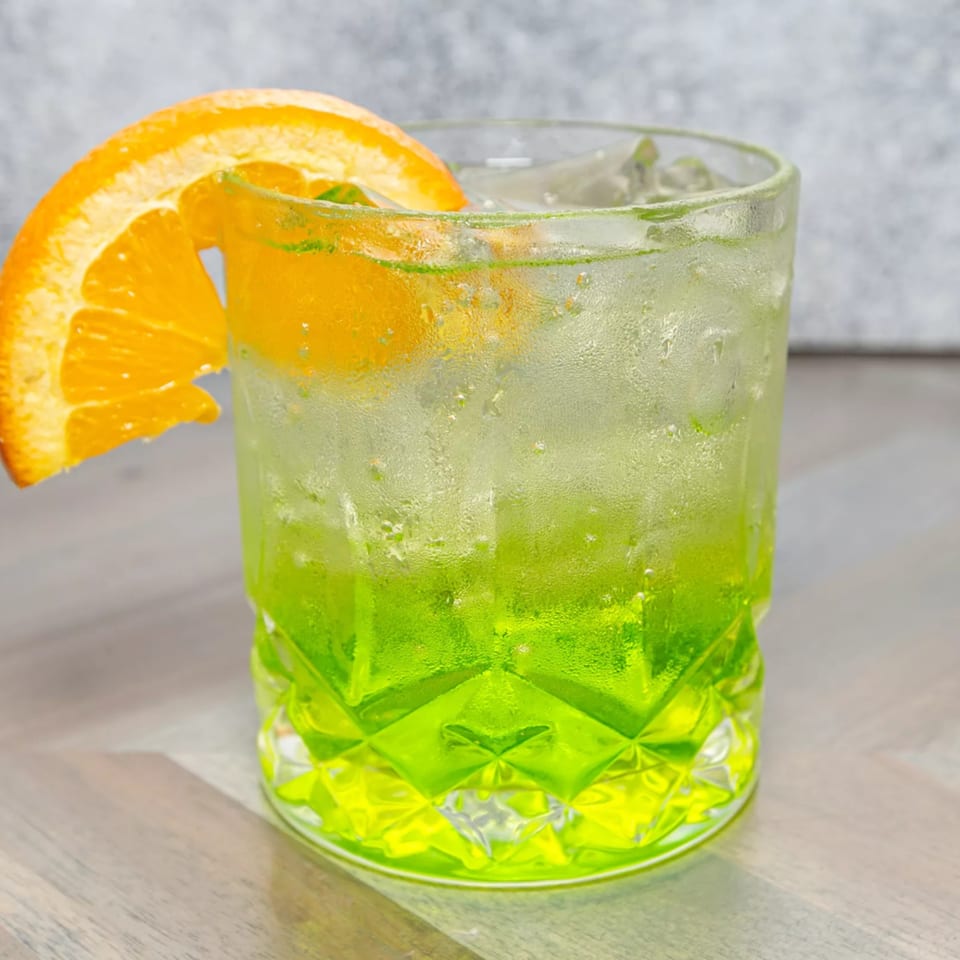 Refreshing green cocktail with ice and an orange slice garnish, on a wooden table.
