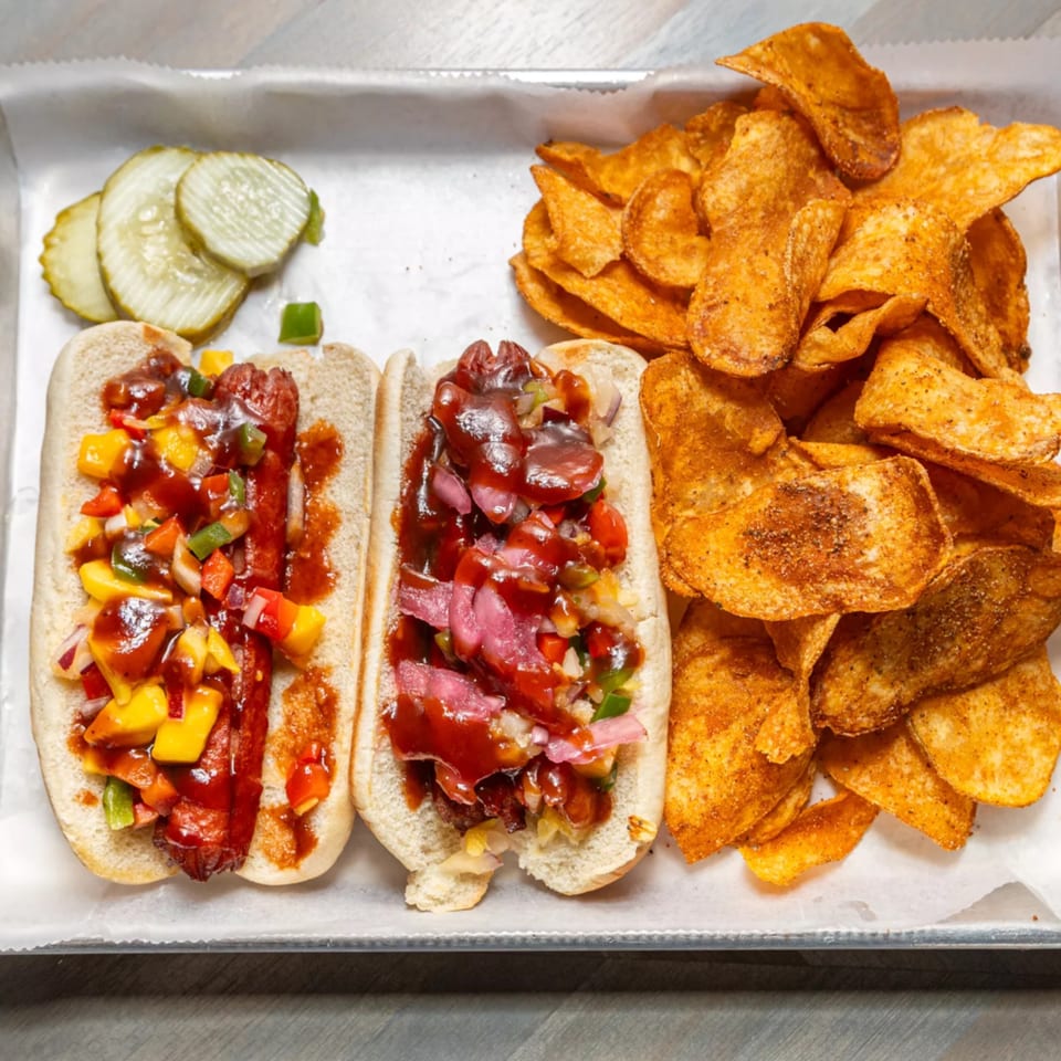 Two hot dogs with colorful toppings and sauce, served with seasoned chips and pickle slices.