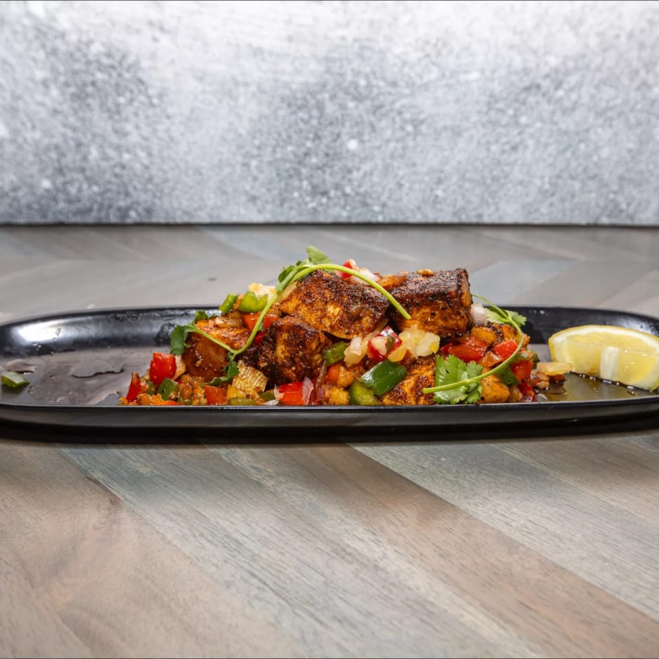 Blackened fish pieces with diced vegetables and cilantro, garnished with a lemon wedge on a black plate.